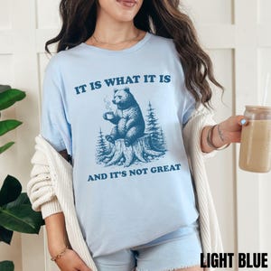 May include: Light blue t-shirt with a graphic of a bear sitting on a tree stump, holding a mug. The text on the shirt reads "IT IS WHAT IT IS" and "AND IT'S NOT GREAT". The shirt is a light blue color.