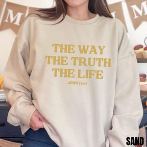 May include: Sand-colored sweatshirt with the text "THE WAY THE TRUTH THE LIFE" in a gold color. The text "JOHN 14:6" is below the main text. The sweatshirt is a neutral color and has a relaxed fit.