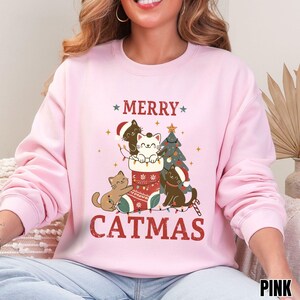 Op de afbeelding: Roze sweatshirt met een "Catmas" ontwerp. De afbeelding toont katten met kerstmutsen, een kerstboom en een kous. De woorden "MERRY" en "CATMAS" zijn gedrukt in een feestelijk lettertype.