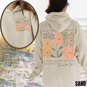 Puede incluir: Una sudadera con capucha beige con un dise&ntilde;o floral y el texto "YOU MATTER" en la parte delantera y "DEAR PERSON BEHIND ME YOU ARE AMAZING, BEAUTIFUL AND ENOUGH Remember That" en la parte trasera.