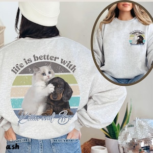 Op de afbeelding: Een grijs sweatshirt met een vintage-stijl grafische afbeelding van een kat en een hond die elkaar knuffelen. De afbeelding zegt "Life is better with Kittie and Bo".