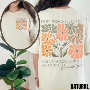 Puede incluir: Una camiseta de color crema con un dise&ntilde;o floral y el texto "Dear person behind me, you are amazing, beautiful and enough. Remember that." en la parte posterior. La parte delantera de la camiseta tiene el texto "You matter" y un dise&ntilde;o floral m&aacute;s peque&ntilde;o.
