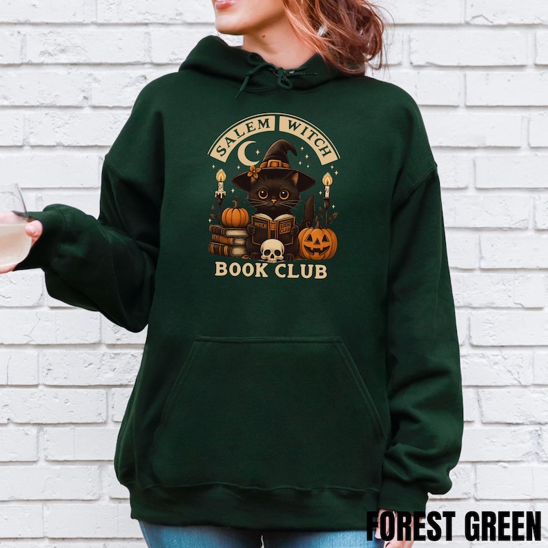 K&ouml;nnte beinhalten: Ein waldgr&uuml;ner Kapuzenpullover mit einem Grafikdruck einer schwarzen Katze mit Hexenhut, die ein Buch liest, umgeben von K&uuml;rbissen, Kerzen, einem Totenkopf und B&uuml;chern. Der Text lautet SALEM WITCH BOOK CLUB in cremefarbener Schrift.