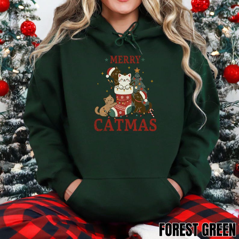 Op de afbeelding: Bosse groene hoodie met een "CATMAS" afbeelding. De afbeelding toont katten met kerstmutsen en een kerstboom. Het woord "MERRY" staat boven de afbeelding. De hoodie heeft een voorzak en een capuchon met trekkoord.