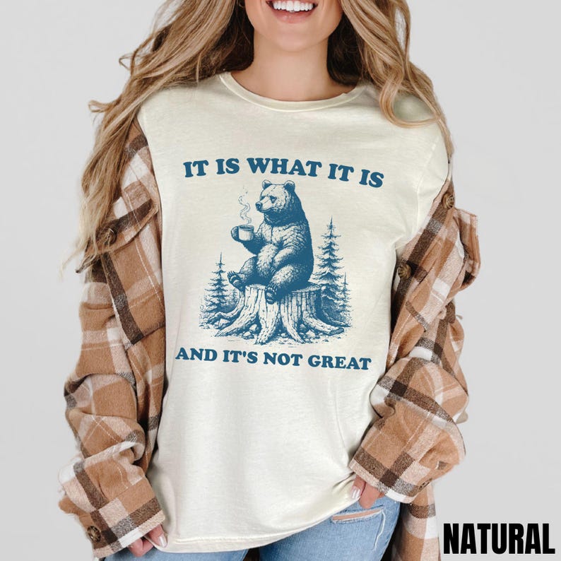 May include: Cream-colored t-shirt with a blue graphic of a bear sitting on a tree stump, holding a mug. The text "IT IS WHAT IT IS" and "AND IT'S NOT GREAT" are above and below the bear illustration. The word "NATURAL" is at the bottom right.