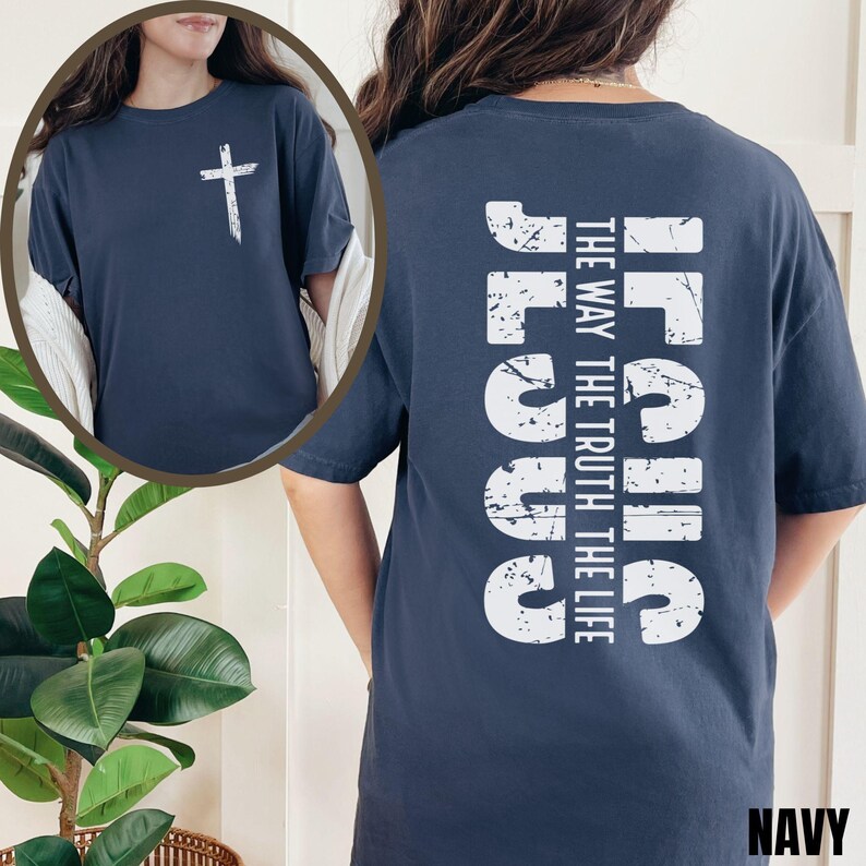 May include: A navy blue t-shirt with a white distressed graphic of the word "Jesus" and the text "The Way The Truth The Life" on the back. The front of the shirt features a white cross.