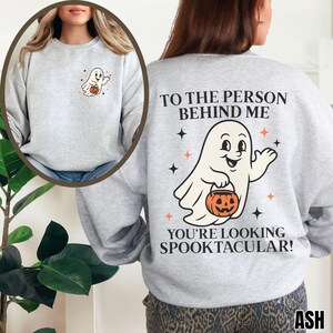Puede incluir: Una sudadera gris claro de cuello redondo con un peque&ntilde;o gr&aacute;fico de fantasma en el pecho izquierdo y un gran estampado en la espalda que dice TO THE PERSON BEHIND ME YOU'RE LOOKING SPOOKTACULAR! con una ilustraci&oacute;n central de un fantasma con calabaza.