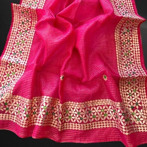 Może przedstawiać: Różowy, przezroczysty sari z bordurą haftowaną złotem i zielenią. Sari ma kratkowany wzór i jest ozdobione małymi, okrągłymi, srebrnymi zdobieniami.