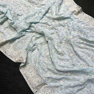 Pu&ograve; includere: Un tessuto azzurro con intricati ricami floreali bianchi. I bordi sono adornati da un bordo dettagliato e piccoli abbellimenti riflettenti. Il materiale delicato &egrave; drappeggiato, mostrando il design dettagliato.