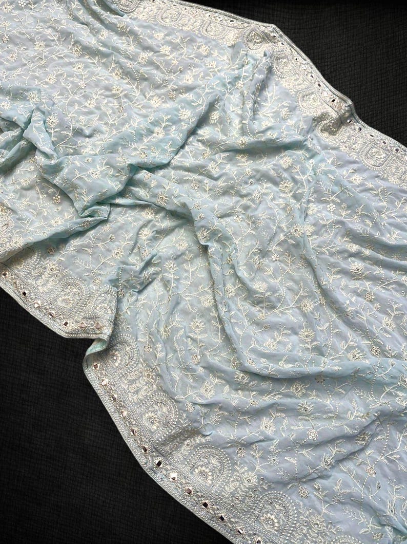 Pu&ograve; includere: Un tessuto azzurro con intricati ricami floreali bianchi. I bordi sono adornati da un bordo dettagliato e piccoli abbellimenti riflettenti. Il materiale delicato &egrave; drappeggiato, mostrando il design dettagliato.