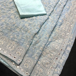 Pu&ograve; includere: Un sari azzurro con intricati ricami floreali bianchi e specchi lungo i bordi. Il sari &egrave; piegato, con un pezzo di tessuto azzurro abbinato sopra. Il tessuto ha un aspetto morbido e fluido.