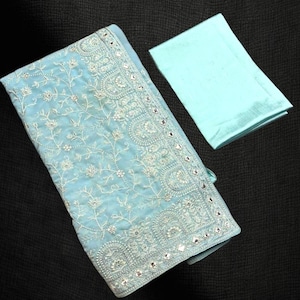 Pu&ograve; includere: Un tessuto ricamato azzurro con intricati motivi floreali e dettagli a specchio. Un quadrato di tessuto azzurro chiaro abbinato &egrave; accanto al tessuto ricamato. Gli articoli sono esposti su uno sfondo scuro.