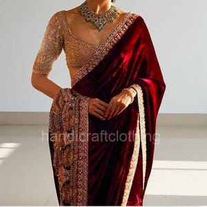May include: A deep red velvet saree featuring a gold embroidered border and a matching gold sequined blouse with elbow-length sleeves. A heavy gold necklace with green stones is worn around the neck. The fabric has a rich, reflective texture.