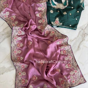 Peut inclure: Un sari mauve avec des broderies florales complexes et des détails de sequins. Le sari est présenté à côté d'un chemisier vert foncé avec une broderie assortie. Le texte "handicraftClothing" est visible.