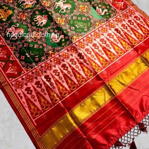 May include: A colourful textile with a detailed pattern in green, red, and gold. The design incorporates repeating geometric shapes and animal motifs, including elephants and horses. The fabric has a silky sheen and a fringed edge. The text "handcraftclothing" is visible.