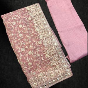 Pu&ograve; includere: Un tessuto ricamato color rosa antico con intricati motivi floreali e un tessuto in tinta unita abbinato. Il tessuto ricamato ha un bordo a specchio. I tessuti sono esposti su una superficie scura.