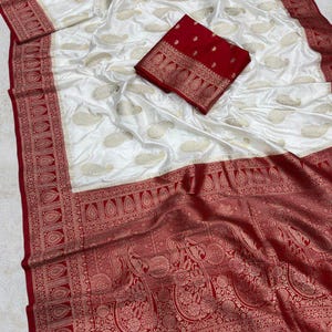 Könnte beinhalten: Weiß-roter Seidensari mit aufwendigen Paisley- und Blumenmustern. Der Sari hat eine weiße Basis mit goldenen Paisley-Motiven und einen roten Rand mit detaillierten Designs. Ein passendes rotes Blusenteil ist enthalten.