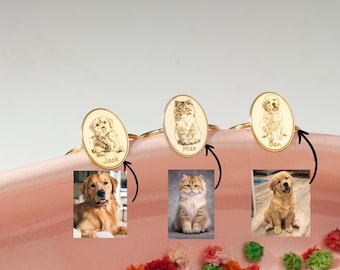 May include: Three gold rings with engraved images of a dog, cat, and dog. The rings are on a pink surface with flowers. The names "Jack", "Max", and "Ben" are engraved on the rings.