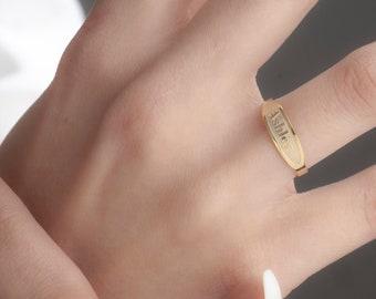 May include: A gold-colored ring with the name "Ashley" engraved on a rectangular plate. The ring is worn on a finger, showcasing its simple yet elegant design. The ring is a piece of jewelry.