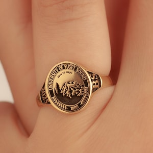 May include: A gold-colored ring with a circular emblem featuring text that reads "UNIVERSITY OF PUGET SOUND" and "2025". The emblem also includes a mountain and tree design. The ring is on a finger.