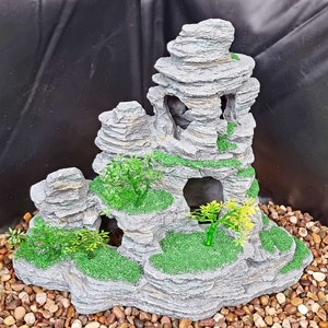 Op de afbeelding: Een grijze stenen rotsformatie met groen mos en kleine groene planten. De rotsformatie heeft verschillende grotten en is ontworpen voor gebruik in een aquarium of terrarium.