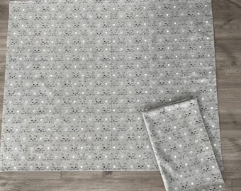 May include: Gray fabric with a repeating pattern of cat faces and small white hearts. The fabric is folded into a rectangular shape, and a larger piece of the same fabric is laid out flat. The cat faces have black whiskers and closed eyes.