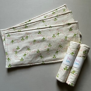 May include: Set of five white burp cloths with a green frog pattern and the words "Baby Care" printed on two rolled up burp cloths.