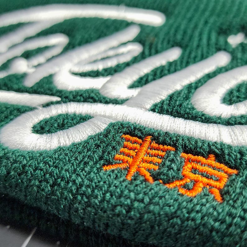 May include: A green beanie with white embroidered lettering and an orange embroidered symbol. The symbol is a stylized Japanese character.
