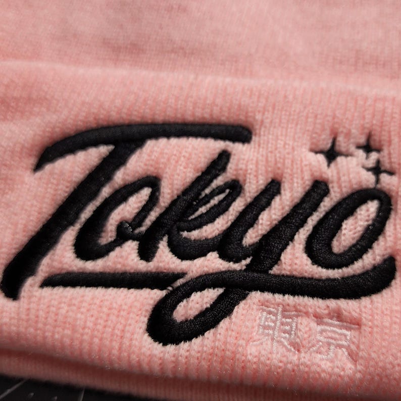 May include: Pink beanie with black embroidered text that says "Tokyo" with a small star design above the "o".