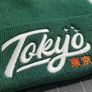 May include: A green beanie with white embroidered text that says "Tokyo" and orange text that says "東京".