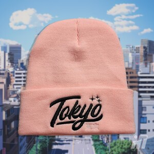 May include: A pink beanie with a black embroidered logo that says "Tokyo" with two stars above the word.