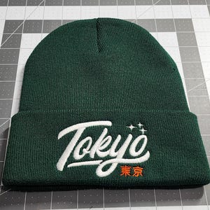 May include: A dark green beanie with a white embroidered "Tokyo" in a stylized font. The word "東京" is embroidered in orange below the word "Tokyo".