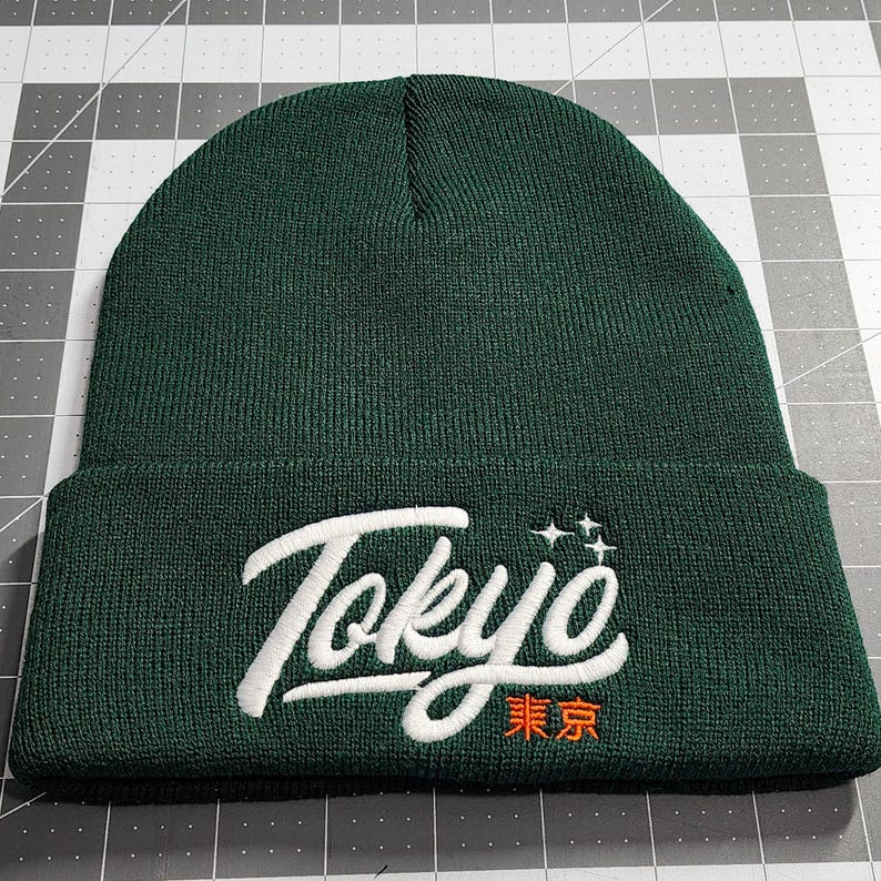 May include: A dark green beanie with a white embroidered "Tokyo" in a stylized font. The word "東京" is embroidered in orange below the word "Tokyo".