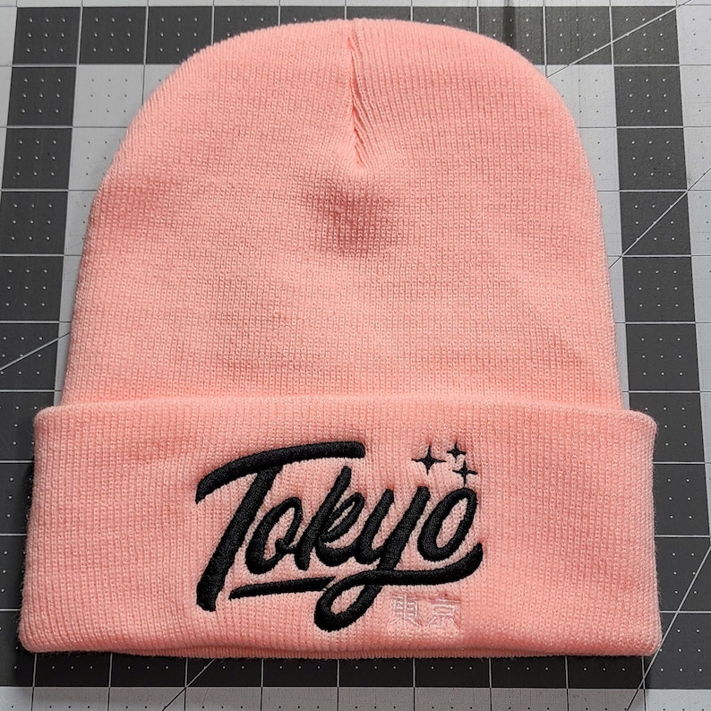 May include: A pink beanie with a black embroidered logo that says "Tokyo" with two stars above the word.