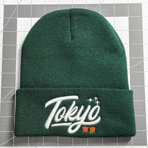 May include: A dark green beanie with a white embroidered logo that says "Tokyo" in a cursive font with a small star design above the word. The word "Tokyo" is also written in Japanese characters in orange below the word.