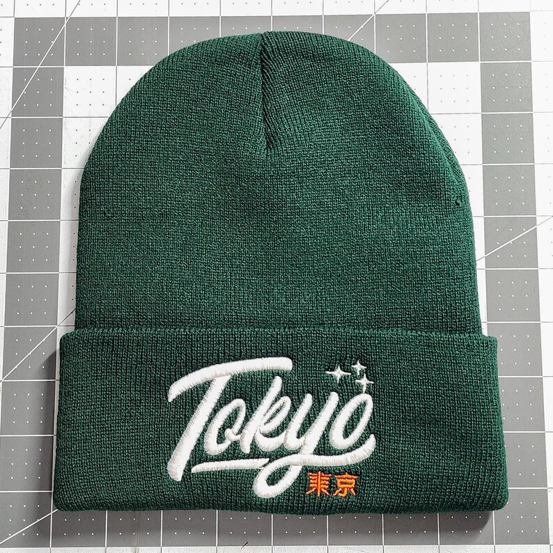 May include: A dark green beanie with a white embroidered logo that says "Tokyo" in a cursive font with a small star design above the word. The word "Tokyo" is also written in Japanese characters in orange below the word.