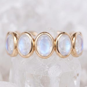 Peut inclure: Une bague en or ornée de cinq pierres de lune ovales taillées en cabochon, disposées en rangée horizontale. Chaque pierre est sertie dans une monture en or lisse, révélant un éclat bleu irisé sur l'anneau en métal doré poli.