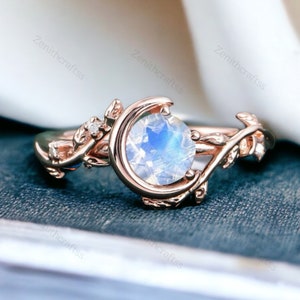 May include: A rose gold ring featuring a crescent moon design and a large, round moonstone. The ring is adorned with delicate leaf accents and small, clear stones.