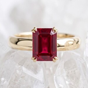 May include: A gold ring featuring a rectangular emerald-cut deep red gemstone set in a four-prong setting. The band is smooth, polished, and rounded, showcasing a classic design that highlights the vibrant color and geometric facets of the center stone.