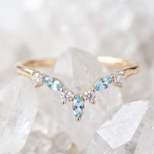 May include: A gold V-shaped ring featuring a central marquise-cut light blue gemstone, flanked by two round-cut clear stones, two additional marquise-cut light blue gemstones, and two round-cut clear stones at the ends of the band.