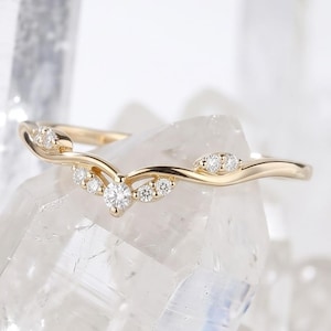 May include: A gold ring featuring a V-shaped band with a central round diamond and smaller accent diamonds set along the curve. The metal band has a polished finish and follows a delicate, contoured silhouette that tapers towards the sides.