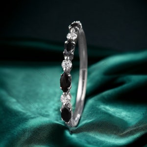 May include: A delicate eternity band ring featuring alternating black and white oval-shaped gemstones. The ring is set in silver and is displayed on a green fabric background.