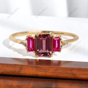 May include: A gold ring featuring a central emerald-cut dark pink gemstone flanked by two smaller rectangular-cut pink stones and two tiny clear triangular side stones. The band is thin and polished gold, showcasing a three-stone design.
