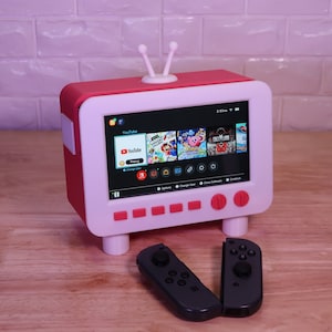 May include: A red and white retro-style television with a screen displaying the YouTube app and other icons. Two black Nintendo Switch Joy-Con controllers are on a wooden surface.