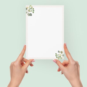 May include: A rectangular white card with a decorative border and watercolor eucalyptus leaf accents in the corners. The card is held by two hands against a light green background.