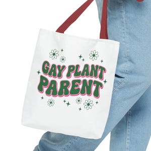 Pode incluir: Bolsa tote branca com alças bordô. A bolsa exibe o texto "GAY PLANT PARENT" em verde e rosa, decorada com motivos de flores e estrelas. A bolsa é segurada por uma pessoa vestindo jeans azul.