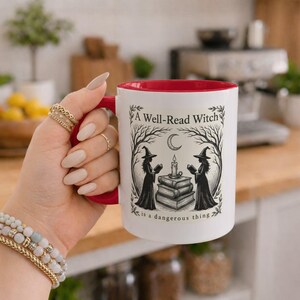 Peut inclure: Un mug en céramique blanc avec un intérieur et une anse rouges, orné d'une illustration à l'encre noire représentant deux sorcières lisant des livres à la lueur d'une bougie sous un croissant de lune, avec le texte A Well-Read Witch is a dangerous thing.