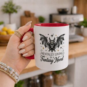 Peut inclure: Un mug en céramique blanc avec un intérieur et une anse rouges, orné d'une illustration en noir et blanc d'une chauve-souris entourée d'étoiles dorées. Le texte "Mentally Living In A Fantasy Novel" est imprimé en noir sous le motif.