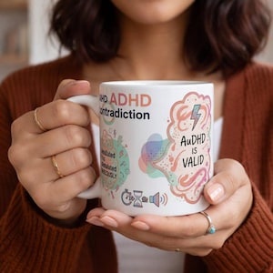 Peut inclure: Mug en céramique blanche avec des graphiques et du texte colorés. La tasse présente les mots "ADHD contradiction" et "AuDHD IS VALID" ainsi que d'autres illustrations. La tasse est tenue par une personne.