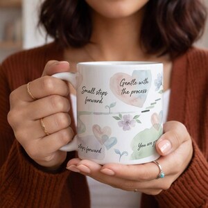 Könnte beinhalten: Weiße Keramiktasse mit dem Text "Small steps forward" und "Gentle with the process" in schwarzer Schrift. Die Tasse hat Aquarellherzen und florale Akzente in Pastellfarben. Die Tasse wird von einer Person gehalten.